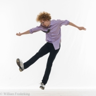 A male presenting light skinned man with a reddish Afro in a purple shirt, jeans and tap shoes is caught mid kick with arms akimbo