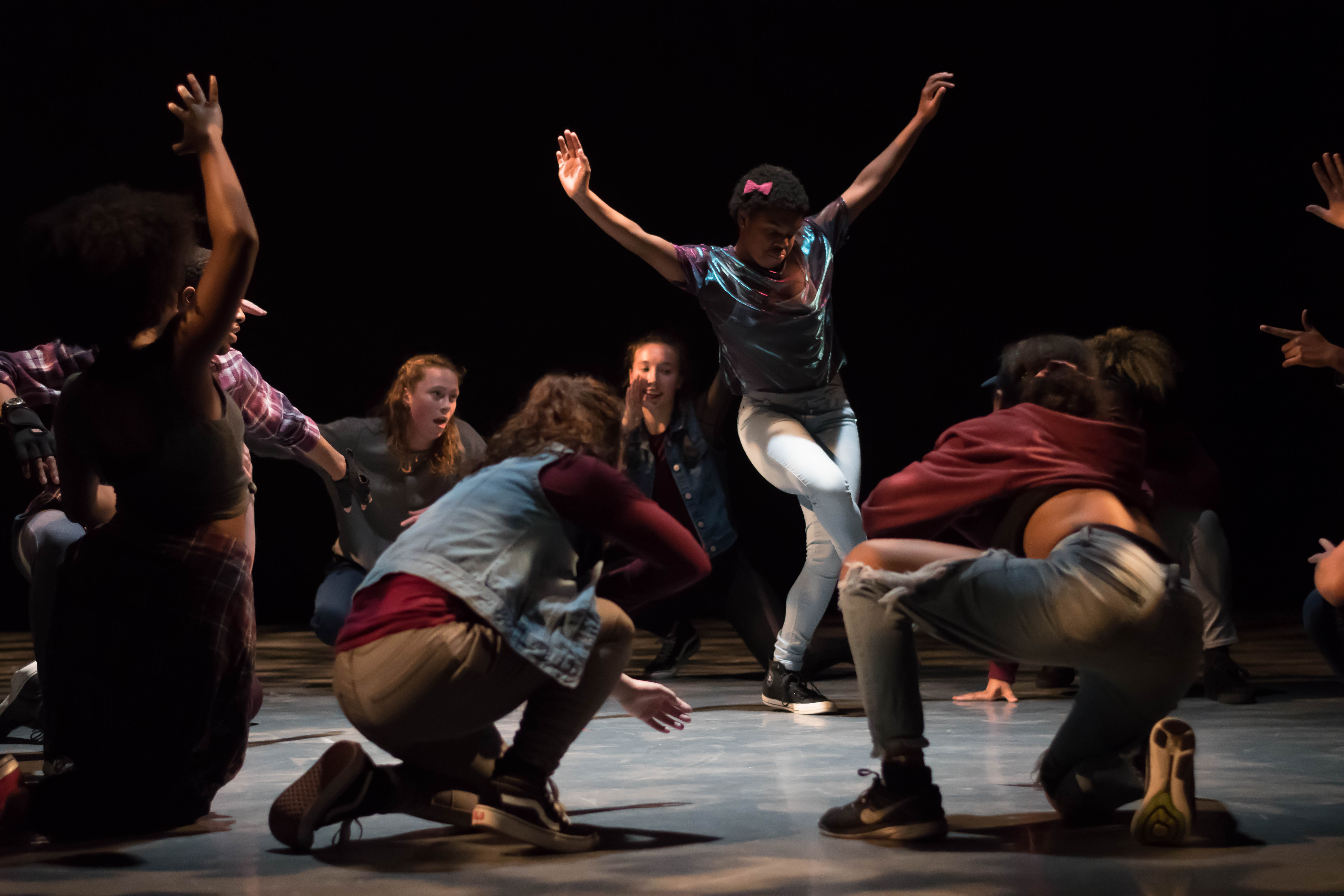 A dancer dances in a middle of a circle of people squatting. Their arms are outstretched. The lighting is dark but the dancers exhibit lots of energy. 