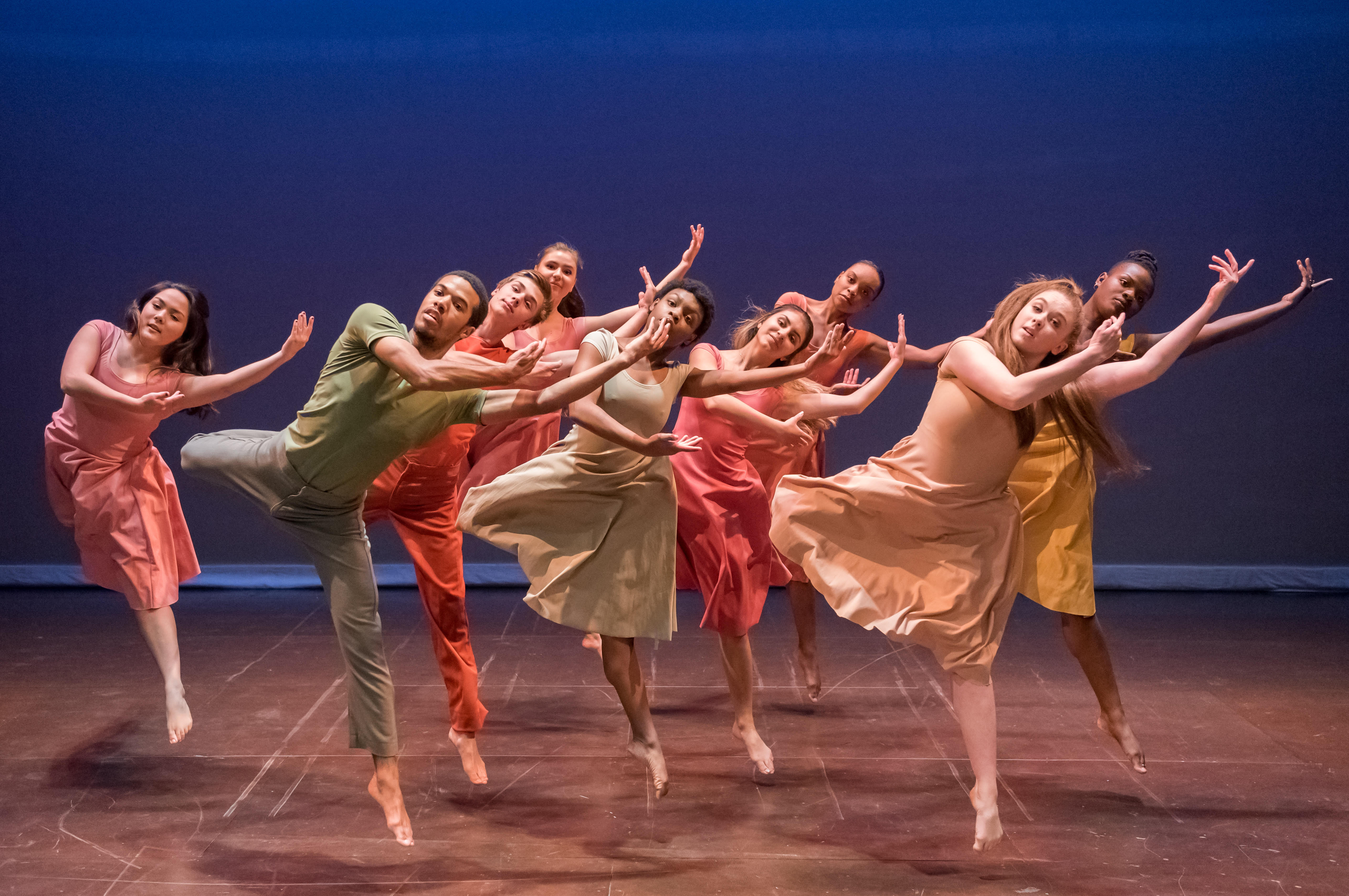 A group of dancers in colorful clothing are arranged in a clump on stage. They are mid jump with their heads and arms towards the right side of the image.