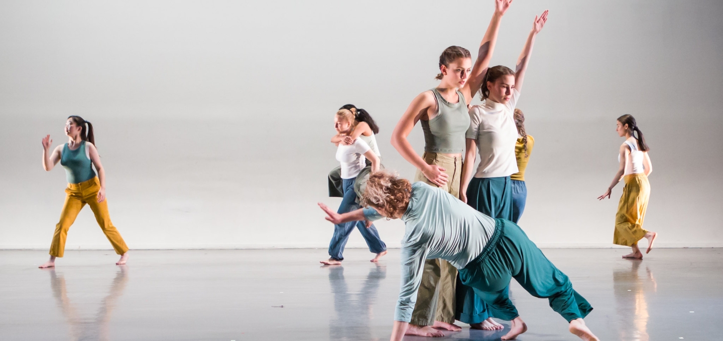 Several dancers on a gray marley floor and white backdrop in  blue, white, and yellow strike a pose in a group. Choreography by Jungeun Kim, photo by Peter Raper