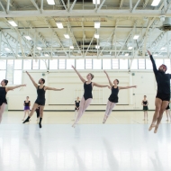 ID: a group of dancers mid jump in a ballet class. They are in a large brightly lit room with light gray marley on the floor. 