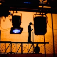 Silhouette of lights, scaffolding, and 2 crew members in conversation against an orange scrim.