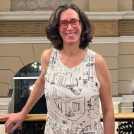 Photo of Michela standing in front of a railing smiling, wearing a black and white patterned dress and red-framed glasses.
