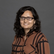 A headshot of Nafisa wearing a brown and black patterned button-up shirt and black-framed glasses.