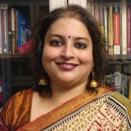 A headshot of Sneha smiling in front of a bookshelf, wearing an orange, green, and red saree.