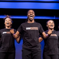 Step Afrika! Nine dancers of African descent dance in a bow shape, arms up at chest level, their T-shirt says Step Afrika!