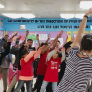 A teacher leads a group of students in a leaning movement with arms overhead.