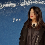 Woman with long brown hair and red lipstick, head turned towards the word "Yes" on blue background with a stream of white dots at the top.