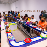 People seated at a conveyor belt touching and looking at art on the belt in a gallery.