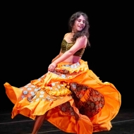 A woman presenting person in a bright orange ruffle skirt twists to show its layers