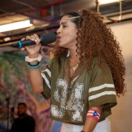 Woman presenting person with long reddish brown curls in a olive green football jersey sings into the mic