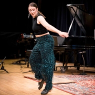 A woman presenting dancer with green sparkle pants tap dances in front of a grand piano