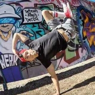 A breakdancer pauses on one arm hold his cap with legs crossed in the air in front of a colorful graffiti wall