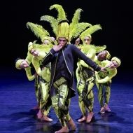A male presenting individual in a lime green hat and blue jacket blows a kiss, surrounded by dancers holding lime green featherssurrounded by 