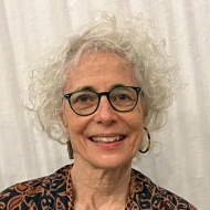 Headshot of a white woman with curly white hair, black rimmed glasses and a dark top.
