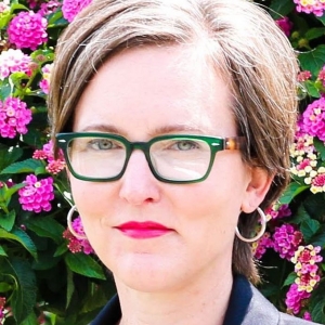 A woman with short brown hair and dark glasses looks at the camera. She is wearing a dark jacket and light top. There are magenta flowers with green leaves in the background.