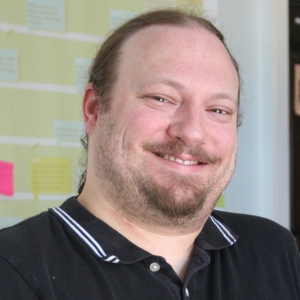 A man with light brown hair and goatee is smiling at the camera. He is wearing a black collared shirt.