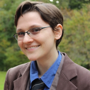 A smiling person with glasses, short brown hair, blue collared shirt, dark tie, and brown jacket.