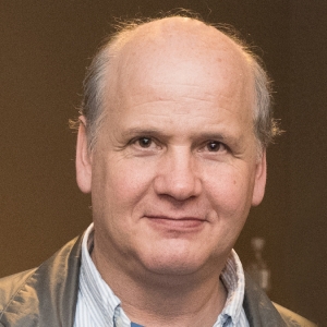 A man with a light collared shirt and grey jacket stares at the camera.