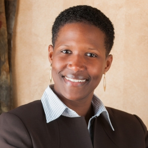 A smiling woman with short black hair, earrings, white collared shirt, and brown jacket.
