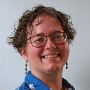 A person with wavy short brown hair, glasses, earrings, and a blue collared shirt.