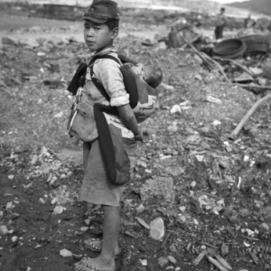A young boy carrying a baby with a destroyed landscape behind them.