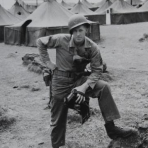 A World War Two soldier standing in front of various military tents with one foot on a large rock.