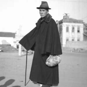 An older person in a hate and long cape is carrying a bag in one hand and a cane in the other.