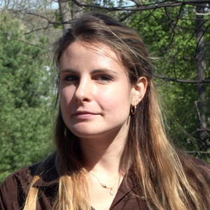 A person with long brown hair wearing a maroon shirt and small necklace with trees in the background.